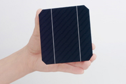 正方形の角を切り落としたような黒い結晶系シリコン太陽電池セルを片手で持つ写真。